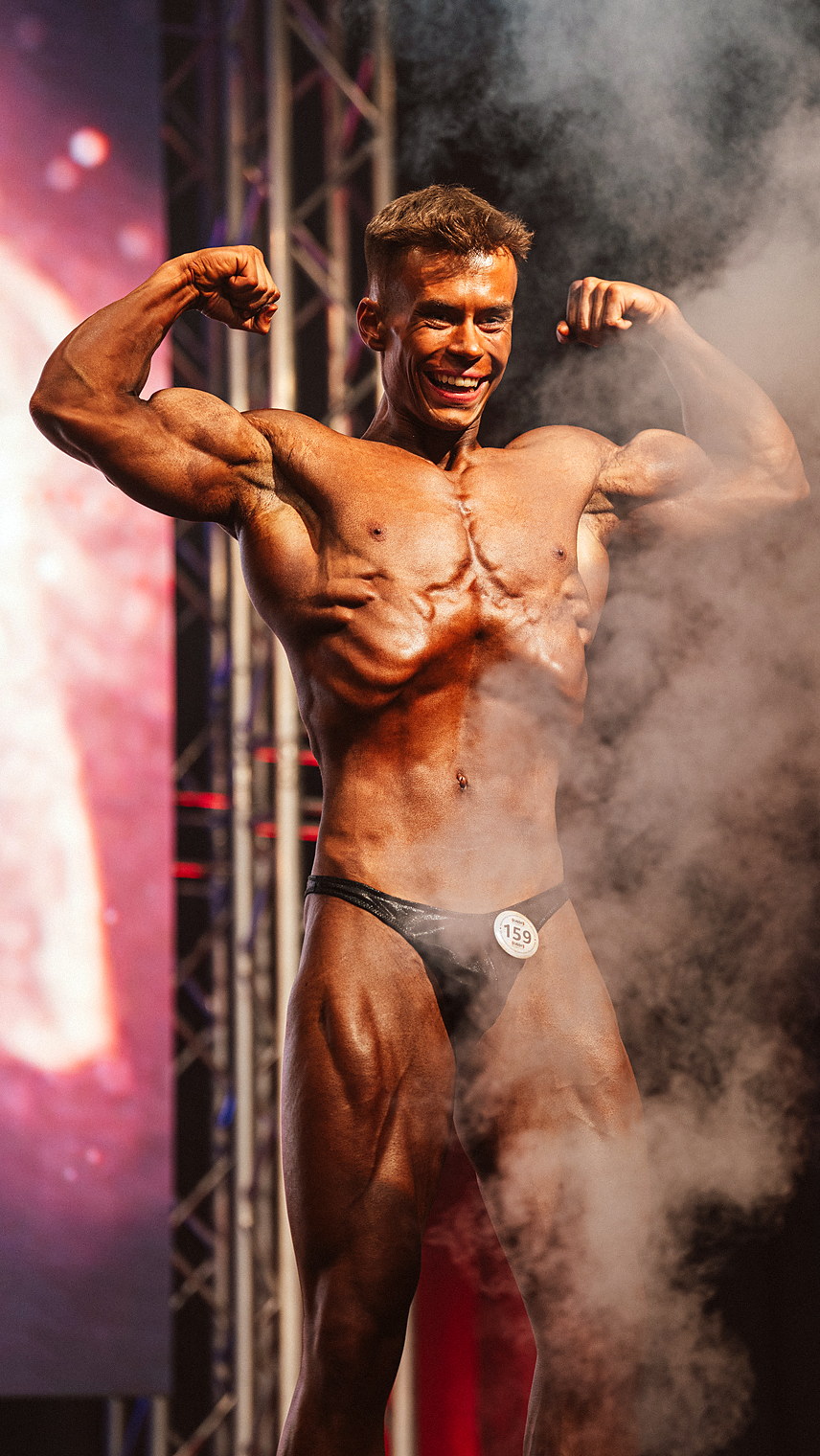 Moritz posiert im Bühnenlicht bei einem Bodybuilding-Wettkampf, beide Arme angewinkelt, Nebel im Hintergrund